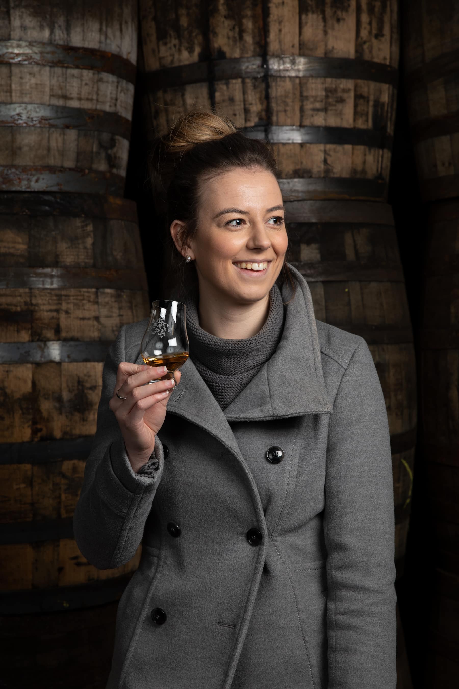 Malt Master Kelsey McKechnie of The Balvenie Whisky holding a glass of whisky in front of stacked wood barrels inside the distillery.