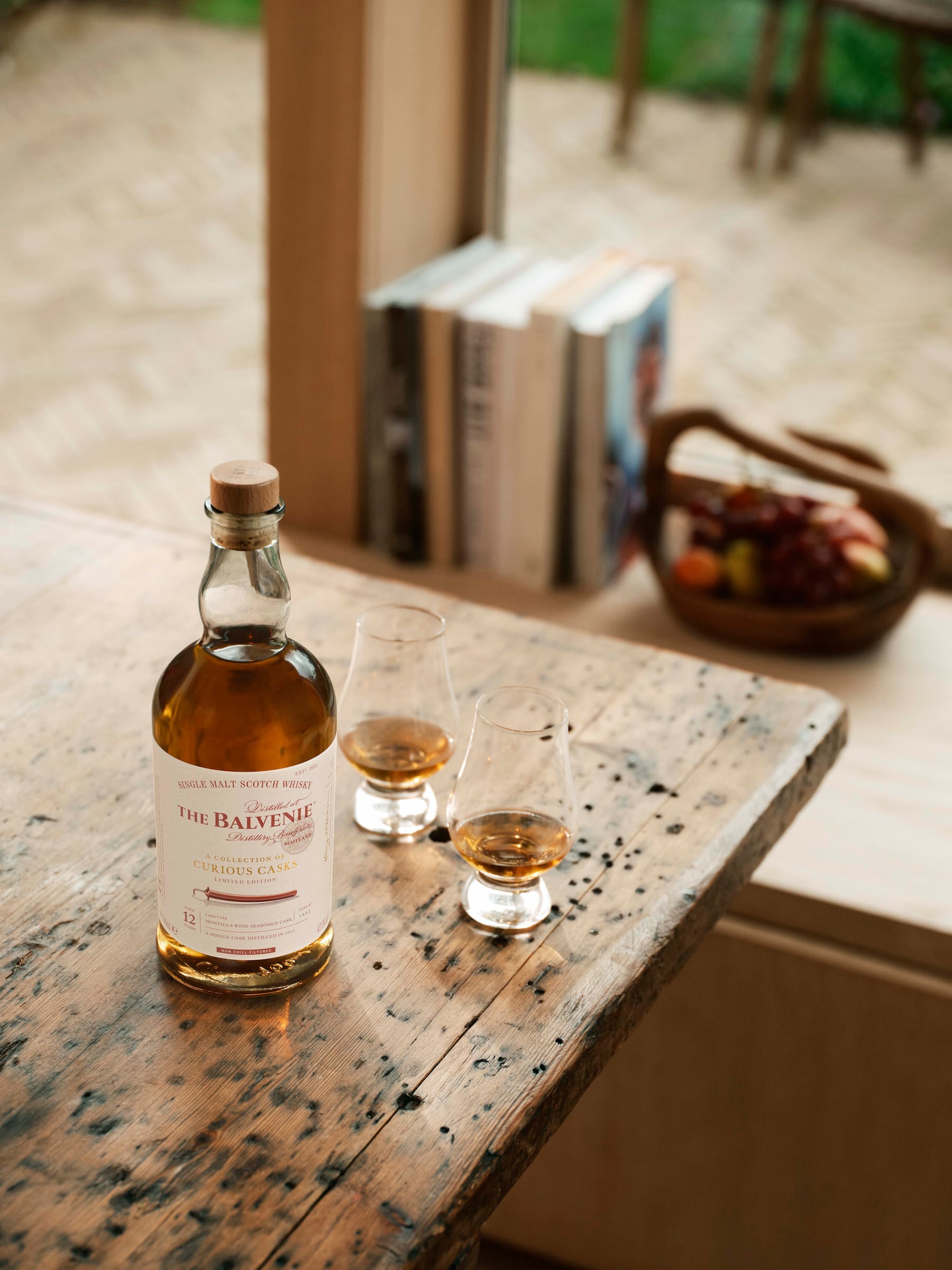 The Balvenie 12 Year Old Montilla Cask Single Malt Scotch Whisky bottle on a rustic wooden table with two filled glasses, set in a cozy room featuring books, fruit basket, and a window view of outdoor seating.
